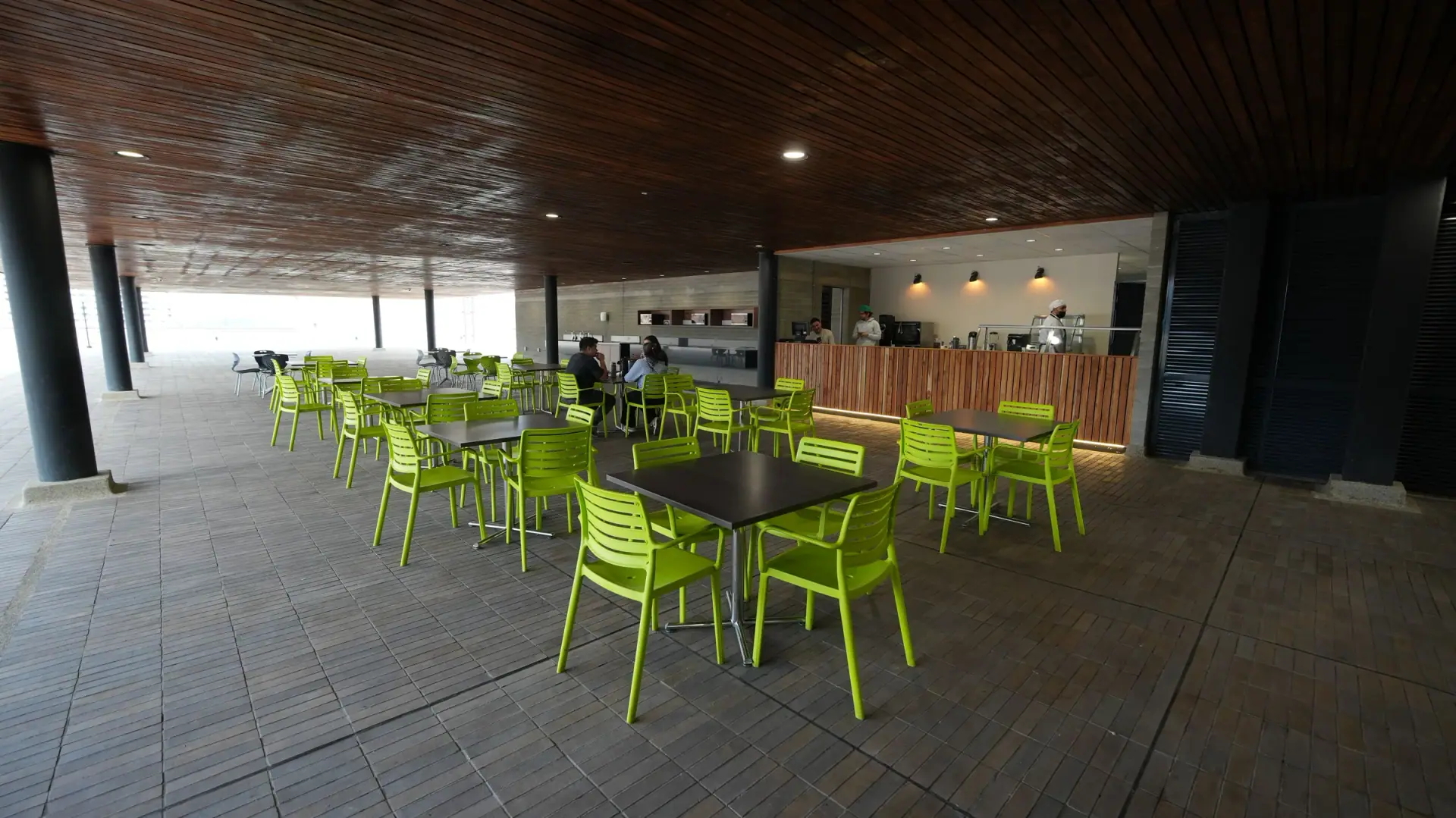 Terraza cubierta de Ayurá Center con mesas negras y sillas verdes junto a la cafetería, bajo un techo de madera