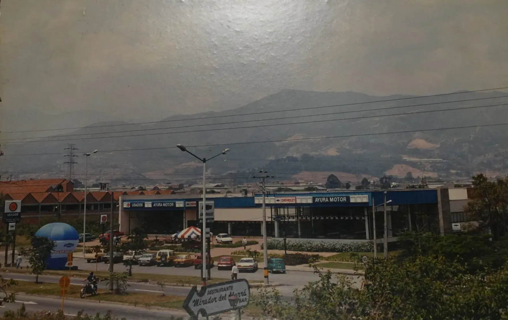 Fotografía de archivo de la antigua sede de Ayurá Motor sobre la avenida Las Vegas, con carros de la época en el parqueadero y las montañas del Valle de Aburrá al fondo