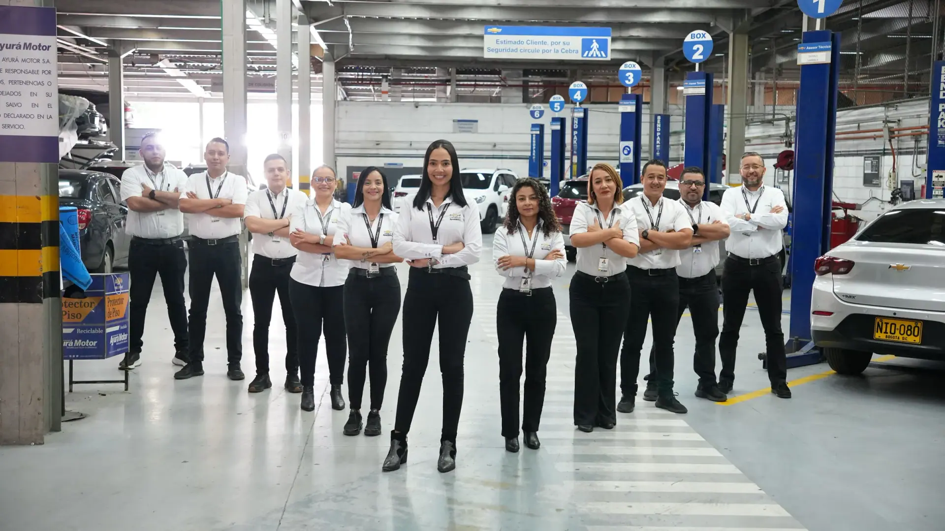 Equipo de posventa de Ayurá Motor, con camisa blanca y brazos cruzados, de pie en el pasillo del taller con los elevadores al fondo