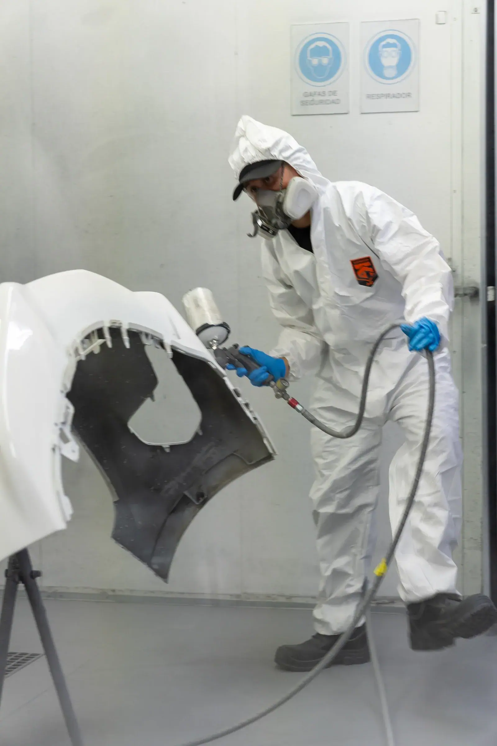 Pintor con traje blanco y respirador aplica pintura a un parachoques en la cabina del centro de colisión